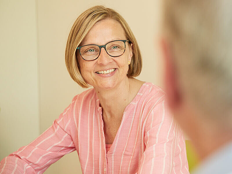 Zwei Personen befinden sich in einer Gesprächssituation. Der Fokus liegt auf einer lächelnden Frau.