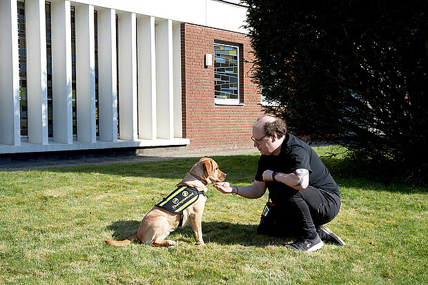 Ein Hundtrainer sitzt mit seinem Hund im Garten der Transitions-Tagesklinik Bochum und erklärt ihm etwas.