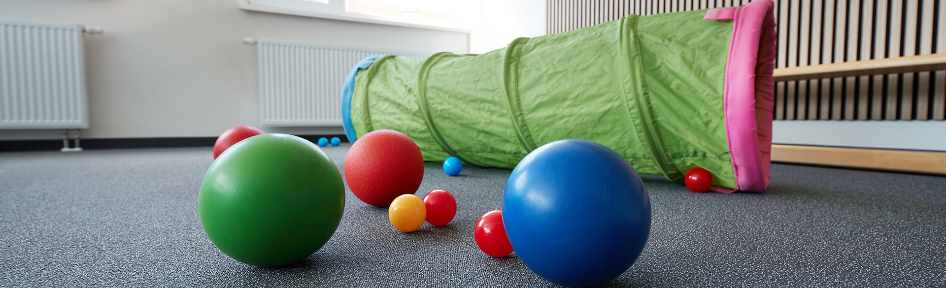 4 bunte Bälle liegen auf einem Teppichboden, im Hintergrund liegt ein Krabbeltunnel.