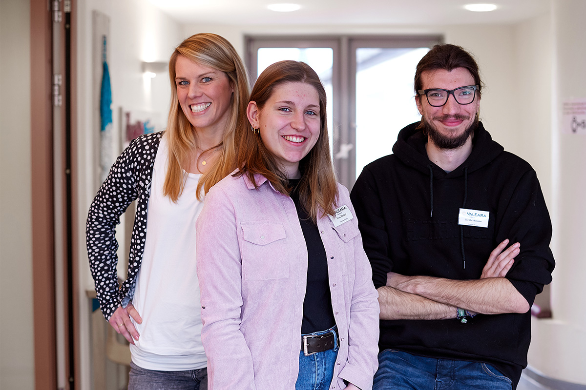 Drei Personen stehen in einem Raum und lächeln in die Kamera.